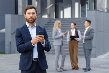 Profesyonel bir işadamı ofisin dışında akıllı telefonuyla uğraşırken meslektaşları arka planda bulanık bir şekilde iş tartışır..