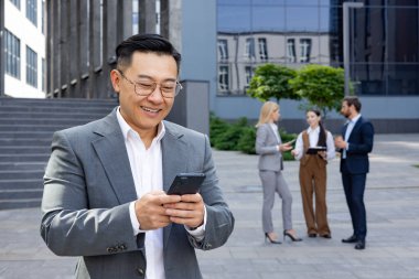 Modern bir ofis binasının arka planında iş arkadaşlarıyla akıllı telefon kullanan iyi giyimli bir iş adamı..