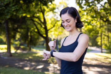 Mutlu genç kadın spor yapıyor ve parkta koşuyor, akıllı saate bakıyor ve eliyle bir evet hareketi gösteriyor..