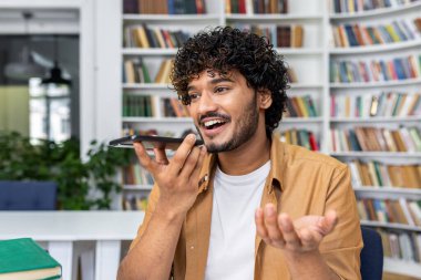 Kıvırcık saçlı neşeli genç adam kütüphane ortamında sesli kumandayı kullanarak akıllı telefonuna konuşuyor..