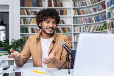 Sıradan bir ortamda yayın yapan, mikrofon ve dizüstü bilgisayarla kitaplıkların önünde canlı olarak tartışan çekici bir podcaster..