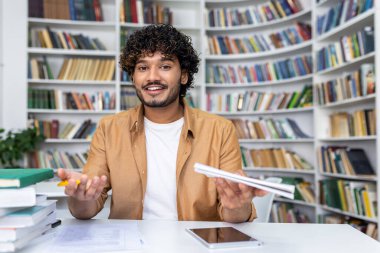 Genç yetişkin, kafası karışmış ve bunalmış bir şekilde bir kütüphane ortamında kitap ve kağıtlarla dolu bir masada oturuyor..