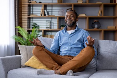 Sakin bir erkek rahat bir nilüfer pozisyonunda bir koltukta oturur, sakin bir oturma odasında meditasyon yapar, sükuneti ve farkındalığı sembolize eder..
