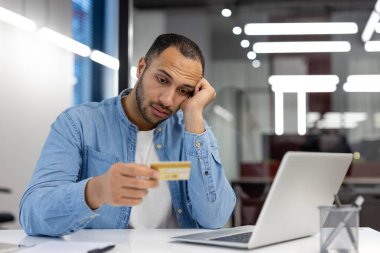 Bir adam kredi kartı tutarken endişeli görünüyor ve dizüstü bilgisayarına bakıyor, muhtemelen mali sorunlarla uğraşıyor..