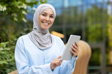 Bir tablet taşıyan mutlu bir Müslüman kadının portresi, arka planda yeşillik ve modern mimariyle dışarıda dikilirken özgüvenini ifade ediyor..