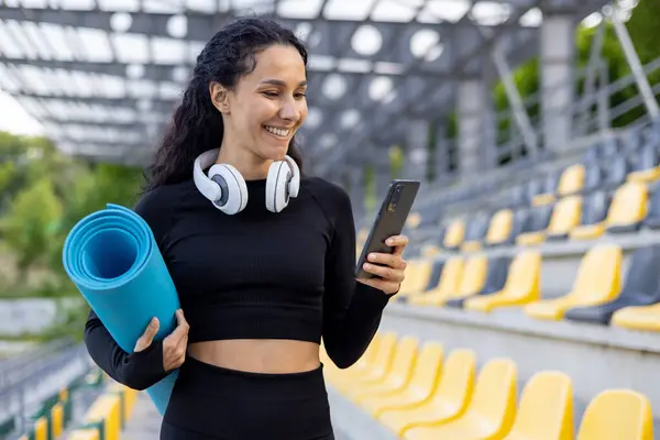 A young athlete with yoga mat and headphones checks her phone at an empty sports stadium, preparing for a workout.