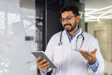 Bir doktor elinde bir tablet tutuyor ve gülümsüyor. Beyaz bir laboratuvar önlüğü ve gözlüğü var. Tablet, içinde bir kişinin resmi olan bir sayfayı açıyor.