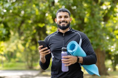 Güneşli, yeşil bir parkta akıllı telefonunu kullanırken elinde yoga minderi ve su şişesi tutan mutlu, formda bir adam..