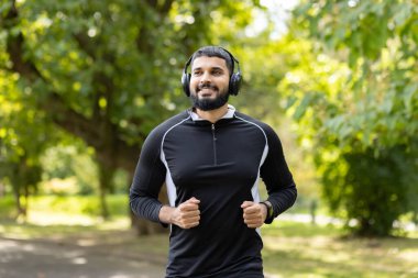 Spor kıyafetli genç bir adam güneşli bir parkta koşuyor, kulaklıklarıyla müzik dinliyor..