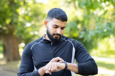 Sportif giyimli bir adam güneşli bir parktaki yeşilliğin ortasında spor takip cihazı kullanıyor. Sağlıklı bir yaşam tarzını gösteriyor..