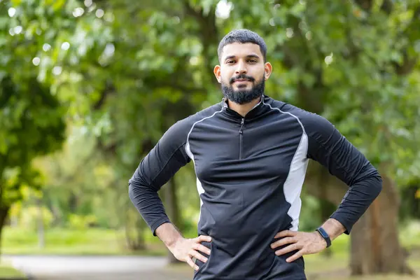 Focused athletic man with hands on hips standing outdoors, wearing sportswear in a park setting, exuding confidence and determination.