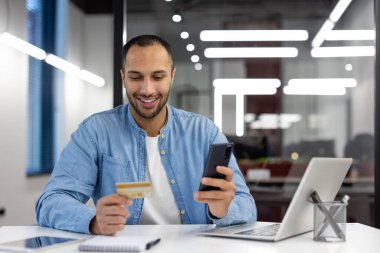 Bir adam kredi kartıyla ödeme yapmak için cep telefonunu kullanırken gülümsüyor. Teknoloji kullanılarak yapılan işlemlerin kolaylığı ve kolaylığı kavramı