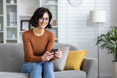 Evdeki bir kanepede rahat rahat oturan rahat bir kadın, akıllı telefonunu iyi aydınlatılmış, rahat bir oturma odasında kullanmakla derinden meşgul..