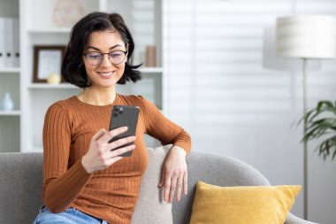 Gözlüklü neşeli genç bir kadın iyi aydınlatılmış bir oturma odasındaki kanepede dinlenirken akıllı telefonuna göz atıyor..