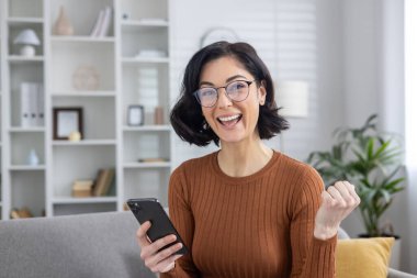 Gözlüklü neşeli genç kadın akıllı telefon tutuyor ve rahat bir oturma odasında muzaffer bir jest yapıyor..