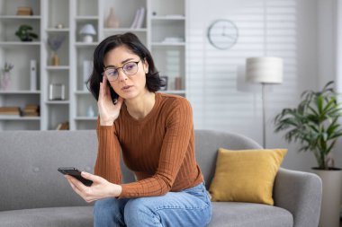 Bir kadın evinde koltuğunda oturmuş, muhtemelen kötü haber aldıktan sonra kafası karışmış bir ifadeyle telefonuna bakıyor..