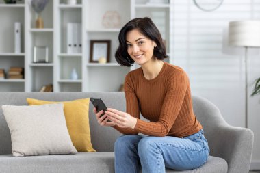 Genç bir kadın akıllı telefonunu kullanırken gülümsüyor, şık, modern bir oturma odasındaki koltukta rahatça oturuyor..