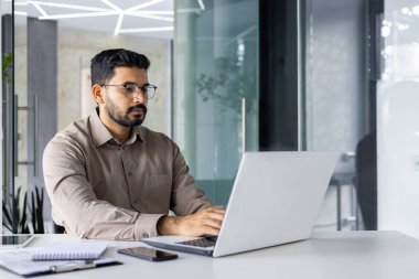 Modern bir ofis ortamında dizüstü bilgisayar kullanırken ciddi ciddi düşünen, şık giyinmiş Hintli bir adam..