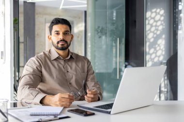 Odaklanmış Hintli işadamı modern ofisinde dizüstü bilgisayarla çalışıyor. Ciddi ve kararlı görünüyor. Profesyonelliği işaret ediyor..