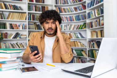 Masasında oturan ve arka planında tasarım kitaplıkları olan kişisel dolabında taşınabilir akıllı telefon tutan aşırı duygusal İslamcı bir adam. Kablosuz dizüstü bilgisayarı, dijital tableti ve kağıtları olan işyeri.