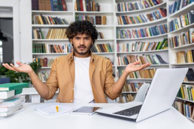 Masasında modern dizüstü bilgisayarı ve kitaplıklarla çevrili kağıtları olan Müslüman bir adam oturuyor. Beyaz gömlekli ve açık kahverengi ceketli bir erkek. İki eli havada, avuç içi kapalı..
