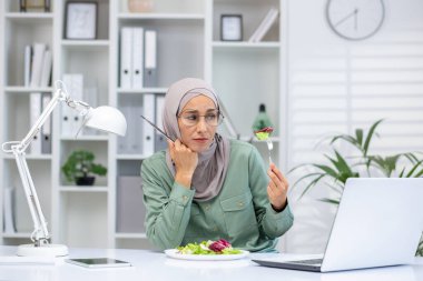 Modern dizüstü bilgisayarlı ve elinde salata yemek için çatal tutan üzgün bir kadının ön görüntüsü. Hüsrana uğramış Arap kadın aşırı yemek yiyor ve ofis ortamında tiksinerek yemek yiyor..