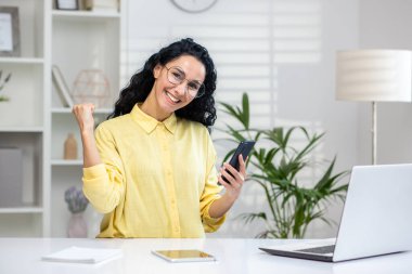 Gözlüklü güzel bir kadının portresi iş yerinde cep telefonuyla oturup olumlu duygularını ifade ediyor. Gülümseyen beyaz kadın kazanan jest yapıyor ve kameraya bakıyor..