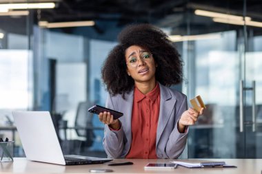 Modern cihazlar ve iş ekipmanlarıyla ofis masasında çalışırken elinde kablosuz akıllı telefon ve kredi kartı tutan Latin bir kadın. Güzel bir kadın çevrimiçi alışveriş yapıyor..