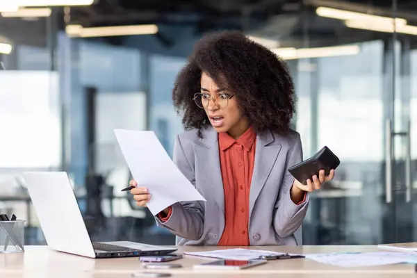 Angry female accountant holding document and calculator in hand while working in office settings. Shocked black woman in glasses sitting in front of modern laptop and performing duties indoors.