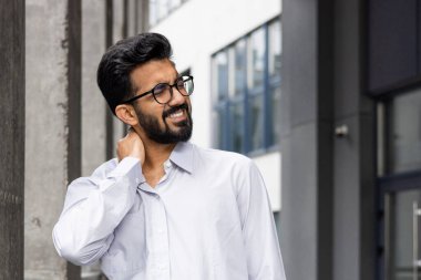 Yorgun genç Hintli erkek ofis çalışanı binanın önünde dikiliyor ve boynunu tutuyor masaj yapıyor, şiddetli acı ve gerilim hissediyor..