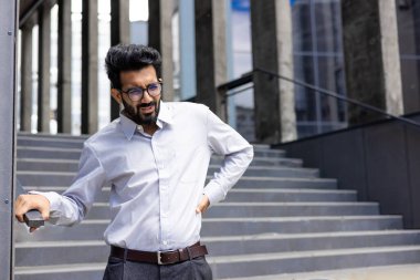 Beyaz gömlekli yorgun genç Hintli adam, binanın yanında duran ofis çalışanı ve arkada el ele tutuşan ağır sırt ağrısı çeken..