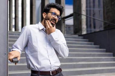 Bir ofis binasının yanında duran Hintli genç adam beyaz bir gömlek ve gözlük takıyor. Çenesini eliyle tutuyor. Şiddetli diş ve çene ağrısı çekiyor..