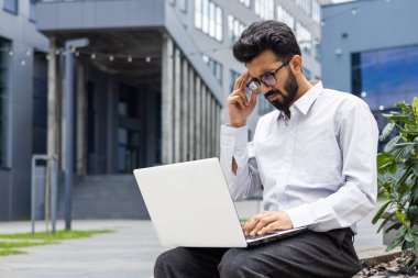 Düşünceli genç Hintli erkek işadamı, öğrenci büro merkezinde bankta oturuyor ve dizüstü bilgisayara endişeli bir şekilde bakıyor..