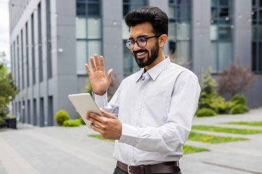 Beyaz gömlekli ve gözlüklü Hintli genç ofis çalışanı internette konuşuyor ve tabletteki video çağrısından selamlıyor..