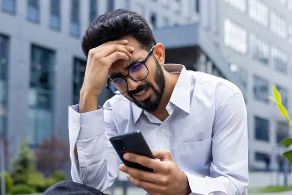 Close-up photo of shocked and upset young Indian man sitting outside holding head with hand and looking at phone screen after receiving bad news.