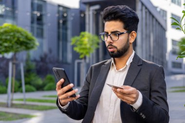 Online hesapta sorun var. Genç endişeli Hintli işadamı dışarıda takım elbiseli, elinde kredi kartıyla duruyor ve telefon ekranına şok olmuş bir şekilde bakıyor..