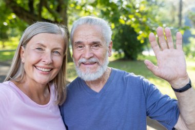 Gülümseyen yaşlı, gri saçlı bir adamın ve bir kadının yakın plan portresi. Parkta bir araya gelip kameraya bakıyorlar ve selamlıyorlar..