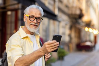 Sırt çantasıyla şehirde yürüyen, gezen, elinde telefon tutan ve kameraya mutlu bir şekilde bakan yaşlı, gri saçlı bir adamın portresi..