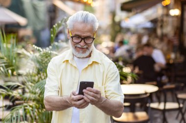 Sarı tişörtlü ve gözlüklü mutlu, kır sakallı bir adam şehirde yürüyor ve gülümseyen bir cep telefonu kullanıyor..