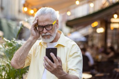 Şok olmuş yaşlı, kır saçlı adam şehir caddesinde duruyor ve endişeli bir şekilde telefon ekranına bakıyor, kötü haberler okuyor, el ele tutuşuyor..