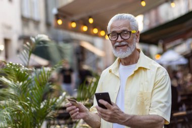 Şehir caddesinde elinde bir kredi kartı ve telefonla kameraya bakıp gülümseyen yaşlı, gri saçlı bir adamın yakın plan portresi..