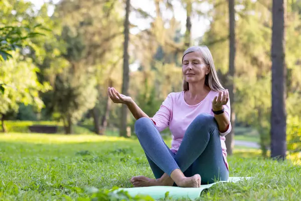 Yaşlı, kır saçlı, rahat bir kadın. Parkta lotus pozisyonunda bir minderin üzerinde oturuyor. Gözleri kapalı ve yoga yaparken meditasyon yapıyor..