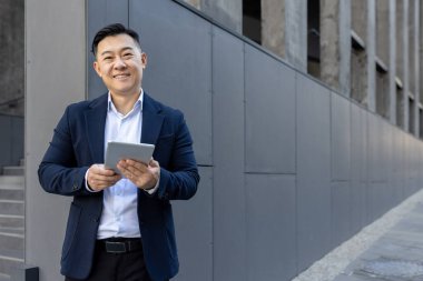 Ofis binasının yanında duran ve elinde tablet olan gülümseyen Asyalı genç bir adamın portresi..