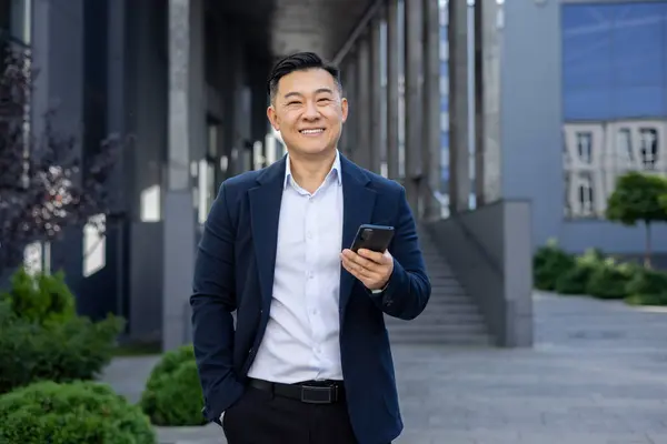 Portrait of Asian successful young Asian man businessman, lawyer, politician in suit standing near office building holding phone and hand in pocket, smiling looking at camera.