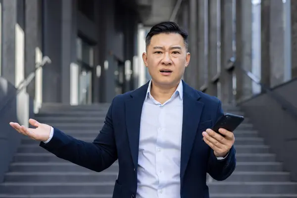 Portrait of a disappointed and shocked young Asian man standing outside a business building, holding a phone, looking upset at the camera and spreading his hands.