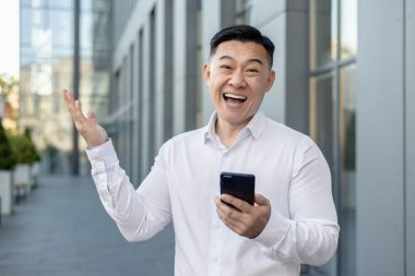 Binanın dışında beyaz gömlekli, elinde telefonla kameraya bakıp şaşırmış ve gülümseyen mutlu bir Asyalı adamın portresi..