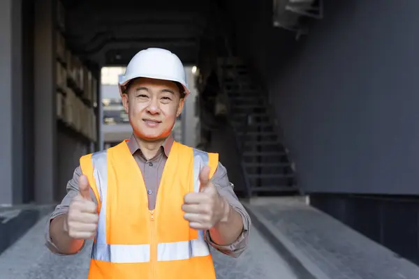 Portrait of a smiling young Asian male construction company worker standing outside in a hard hat and vest, smiling at the camera and showing the super sign with his fingers.