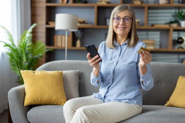 Evdeki bir kanepede rahatça otururken online alışveriş için kredi kartı ve akıllı telefon tutan bir kadının modern tüketim konsepti.