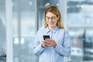 Gözlüklü neşeli bir iş kadını modern ofis ortamında akıllı telefonunu kullanır, profesyonellik ve kendine güven yayar..