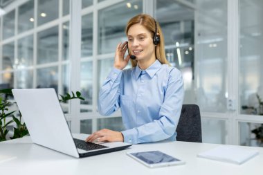 Odaklanmış, mavi gömlekli, kulaklıklı, parlak bir ofis çalışma alanında laptopuyla çalışan profesyonel bir kadın..